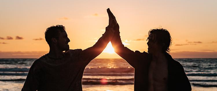 Two friends high-five each other in silhouette at sunset by a beach.