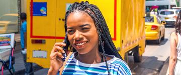 Una mujer con trenzas hablando por teléfono por la calle con un camión amarillo detrás.