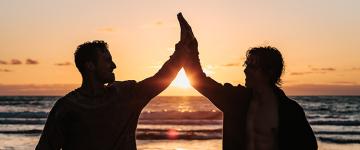Two friends high-five each other in silhouette at sunset by a beach.