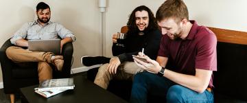 Three friends talk to each other while sitting down. One person has a laptop to look up information, one drinks coffee, and the third takes notes on a notepad.  